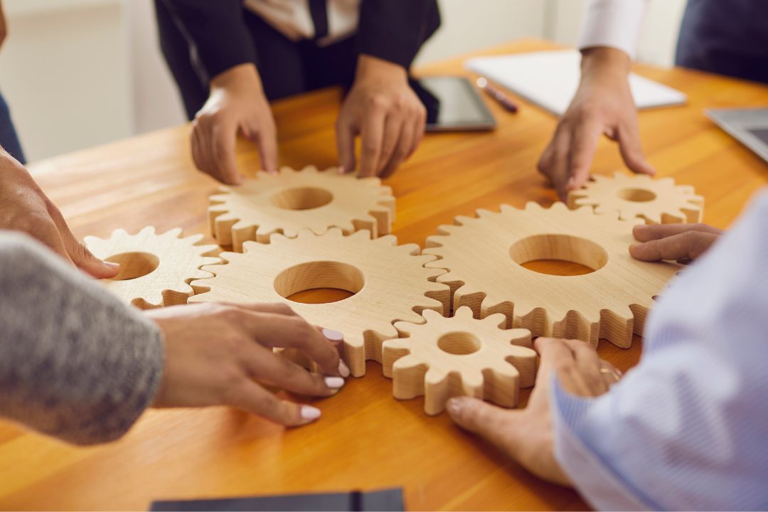 Hands of a diverse business team assembling large wooden gears on a table, symbolizing teamwork, delegation, and business efficiency