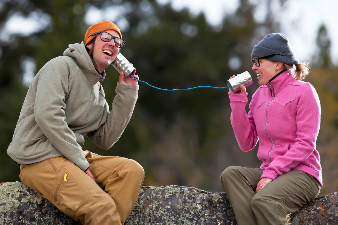 Man and woman sitting outdoors laughing while talking through tin can phones – symbolizing playful communication and listening
