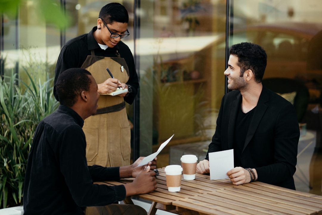 A smiling server in an apron takes an order from two professionally dressed customers sitting outside at a café table, all three engaged and enjoying the moment.