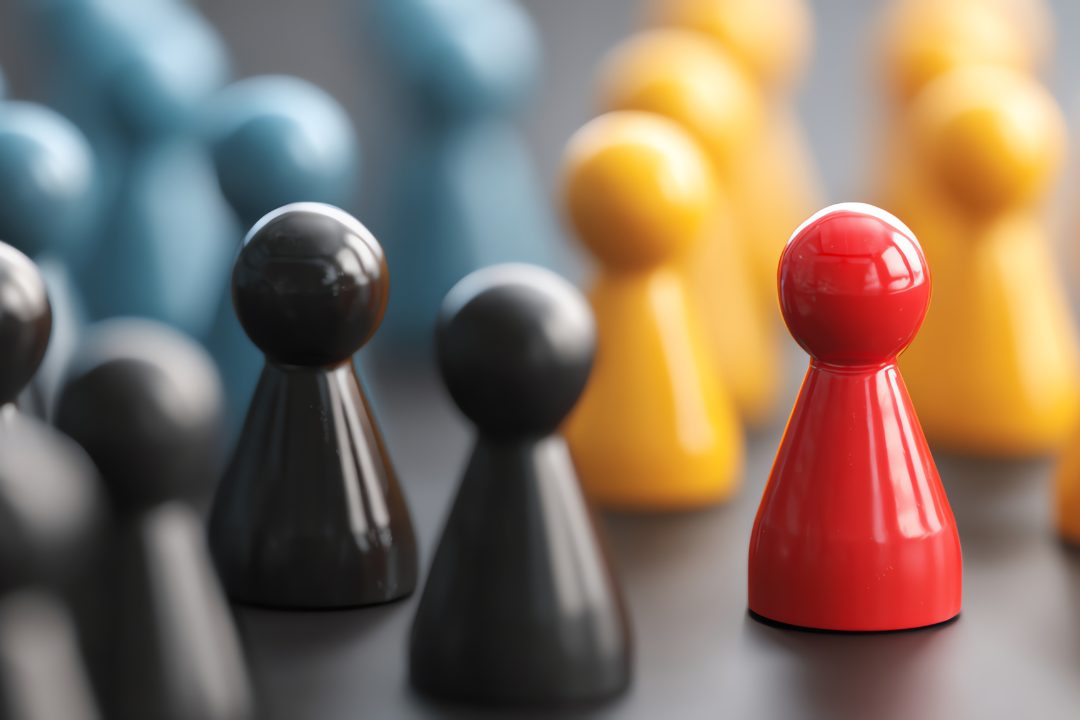 Red game piece standing out from a crowd of black, yellow, and blue pieces, symbolizing differentiation, individuality, and strategic focus in leadership and business.